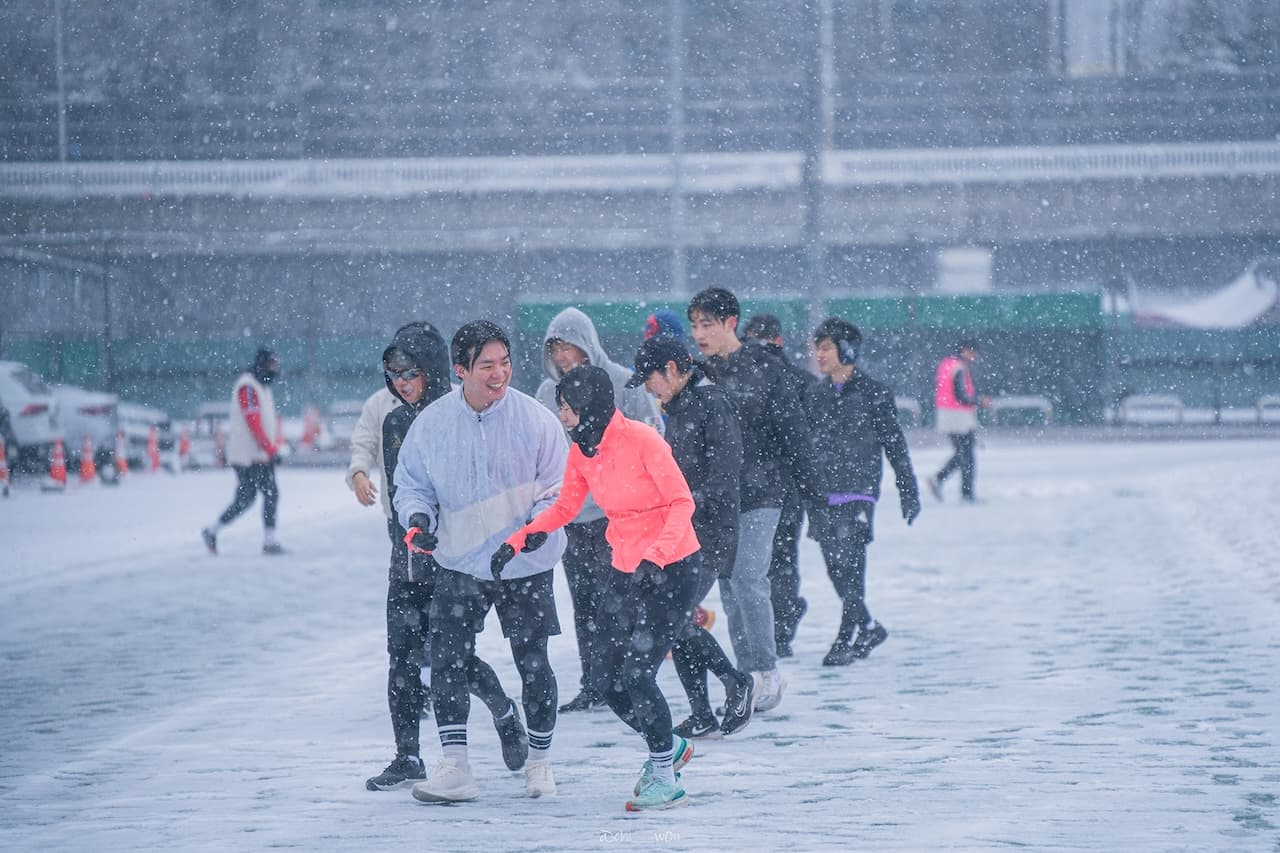 눈이 펑펑 내리는 날, 여러 명의 사람들이 겨울 운동복을 입고 야외에서 함께 달리며 즐거워하는 모습