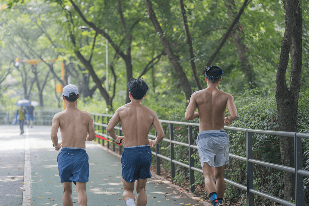 상의를 탈의한 세 명의 남성이 가이드 러닝을 하며 녹음이 우거진 공원 길을 달리고 있는 뒷모습